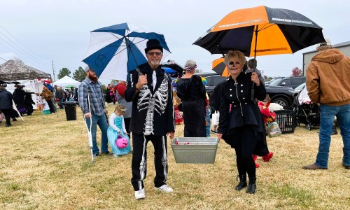 A couple in costumes holding umbrellas at an outdoor event, surrounded by colorful decorations and people.