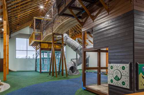 Indoor playground featuring a wooden structure, slide, climbing area, and colorful flooring.