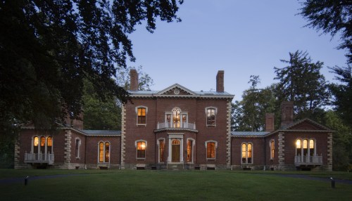 A historic brick mansion with large windows, surrounded by trees, illuminated at dusk.