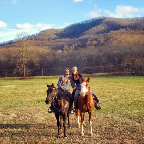 Two people on horseback in a grassy field, with mountains and trees in the background under a clear blue sky.