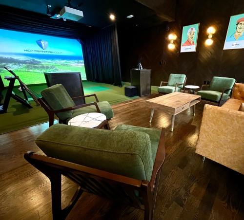 A cozy indoor golf simulator room with green chairs, a wooden table, and a large screen displaying a golf course.