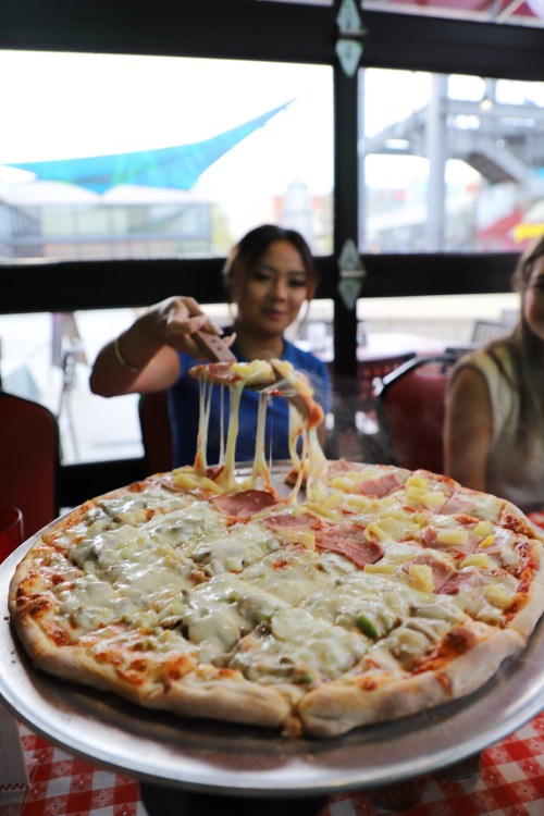 A person serves a large pizza with melted cheese and toppings, sitting at a table in a casual dining setting.