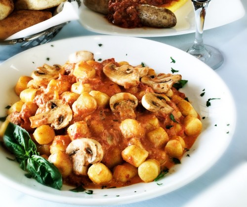 A bowl of gnocchi in a creamy tomato sauce, topped with mushrooms and fresh basil, served with a side of sausage.