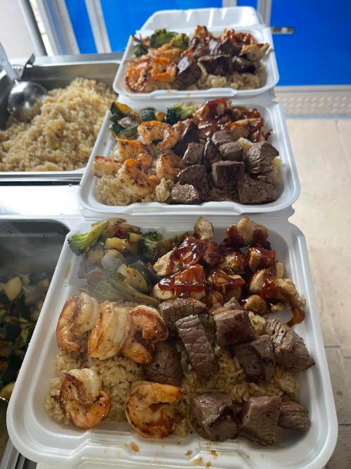 Three takeout containers stacked, each filled with rice, grilled shrimp, beef, and mixed vegetables, drizzled with sauce.