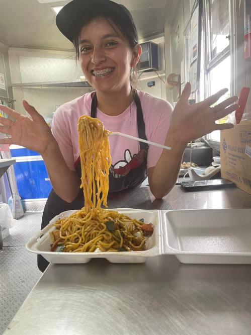 A smiling young woman in a pink shirt holds up a large serving of noodles from a takeout container.