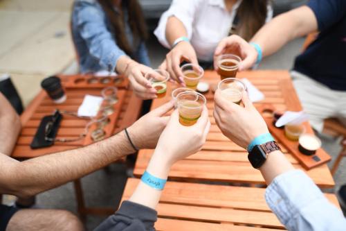 A group of people toasting with small cups of beer at a wooden table, wearing wristbands.