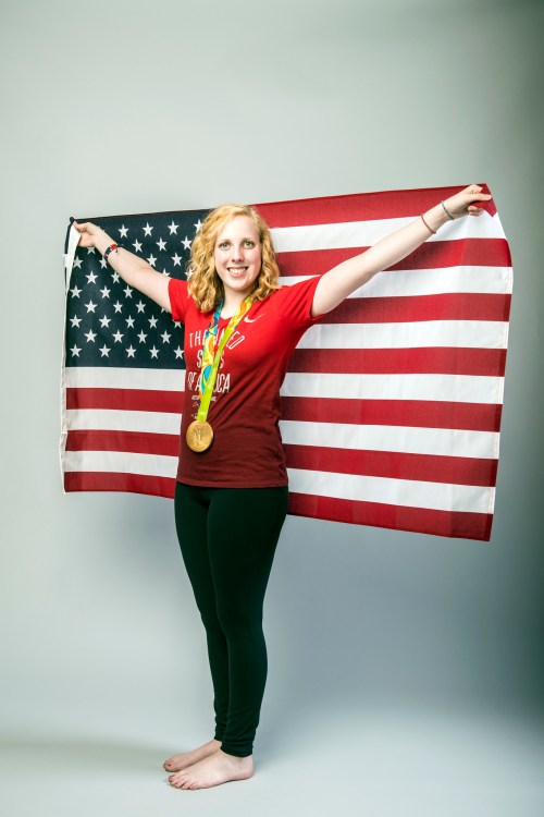 A smiling athlete holds the American flag and displays a gold medal, wearing a red shirt and black leggings.