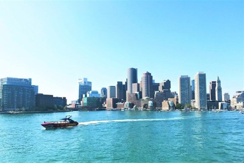 A boat glides across a blue harbor with a skyline of tall buildings under a clear sky.