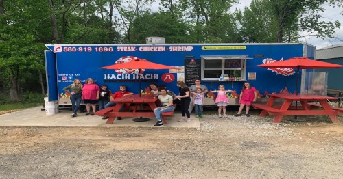 A group of people, including children, poses outside a food truck named "Hachi Hachi" with picnic tables nearby.