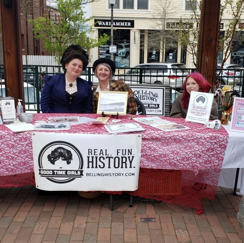 Two women in historical costumes stand behind a table with a red tablecloth, promoting "Good Time Girls" history.