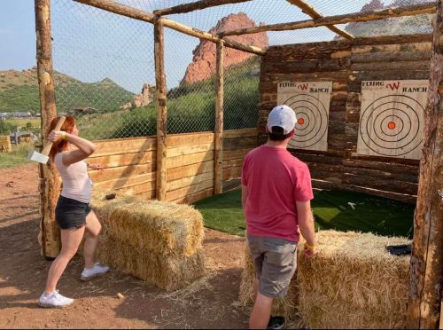 A woman throws a throwing axe at a target while a man watches, surrounded by wooden structures and hay bales.