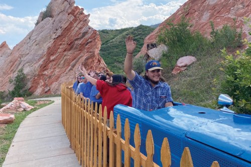 A group of people waves from a colorful train ride near red rock formations and greenery.