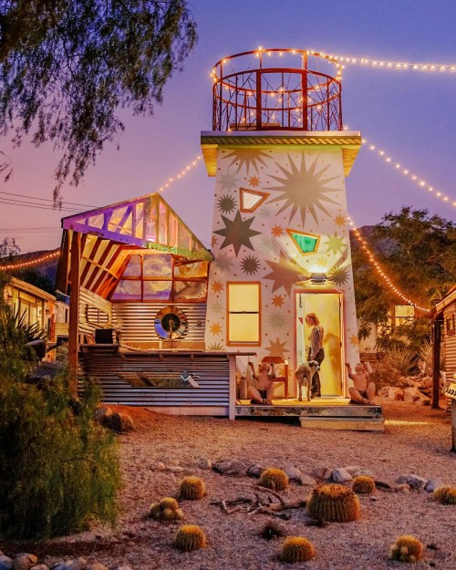 A colorful lighthouse-style building with string lights, surrounded by desert plants and a person with a dog at the entrance.