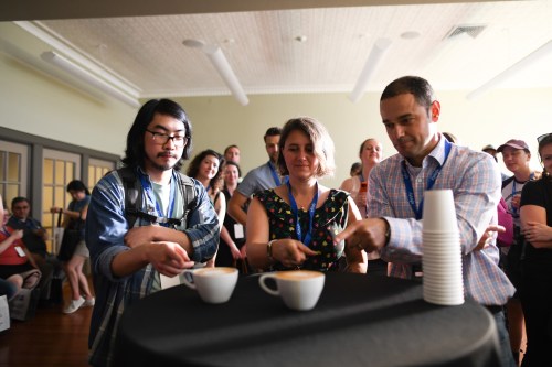Three people participate in a coffee tasting event, each holding a cup while others watch in the background.