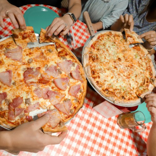 Two pizzas on a checkered tablecloth, with hands reaching in to grab slices. One pizza has ham, the other has vegetables.