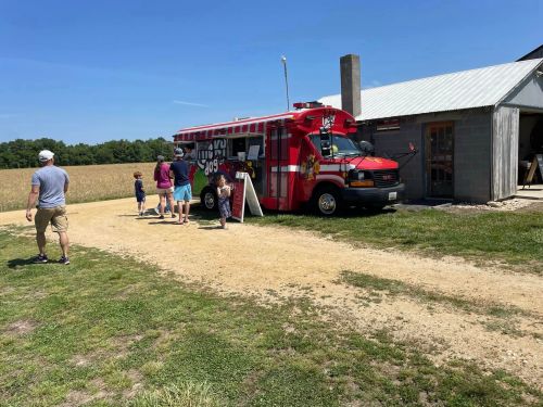 A colorful food truck parked near a field, with people walking towards it on a sunny day.