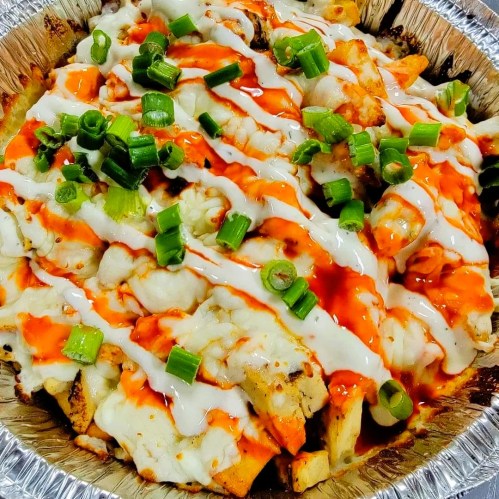 A foil container filled with loaded fries topped with cheese, green onions, and drizzles of hot sauce and ranch dressing.