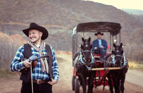 A cowboy in a plaid shirt holds a rifle, smiling, while another cowboy drives a horse-drawn carriage in the background.