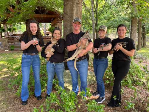 Five women in black shirts hold small animals, smiling in a green outdoor setting with trees and a pavilion.