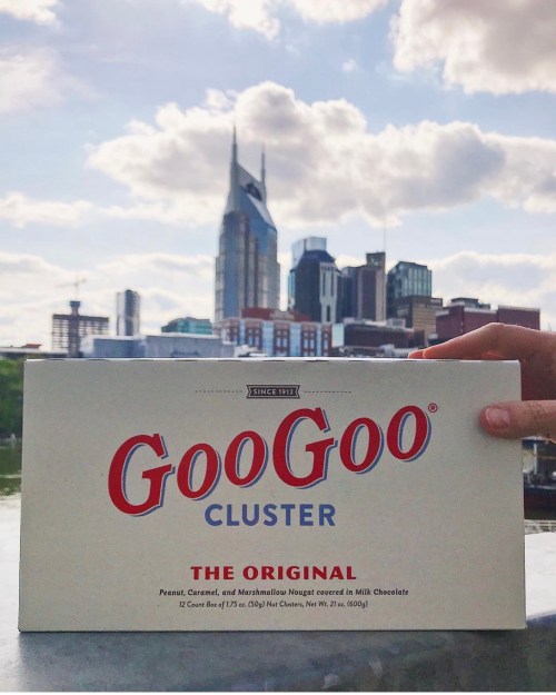A hand holds a box of GooGoo Clusters with a city skyline in the background, featuring tall buildings and clouds.