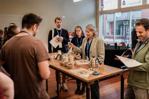 A group of people attentively watches a woman demonstrate coffee brewing techniques at a workshop.
