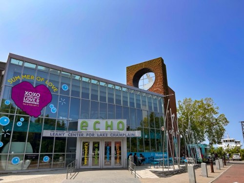 A modern building with large glass windows, featuring colorful signage for the Echo Leahy Center and a sculpture nearby.