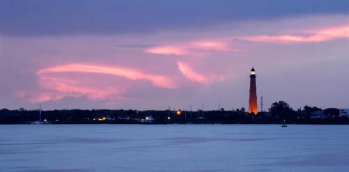 A lighthouse stands against a colorful sunset, reflecting on calm waters with distant lights and clouds in the sky.