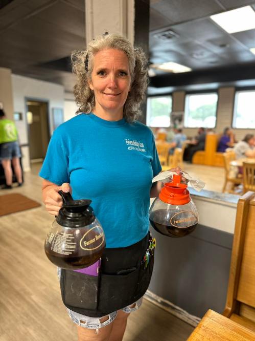 A smiling waitress in a blue shirt holds two coffee pots in a casual dining setting.