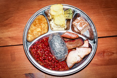 A metal tray with various foods: baked potato, beans, meat slices, applesauce, and a dessert square.