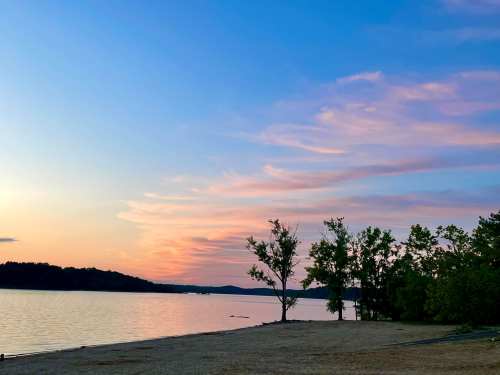 A serene lakeside view at sunset, featuring soft clouds and silhouettes of trees along the shore.