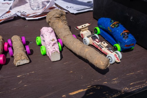 A collection of handmade toy cars, including a pink one, a blue one, and a stick-shaped car, on a wooden table.