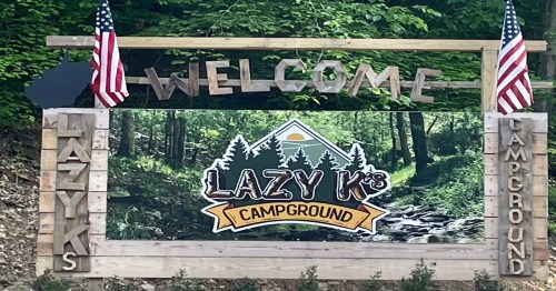 Wooden sign welcoming visitors to Lazy K's Campground, featuring trees, a mountain, and American flags.