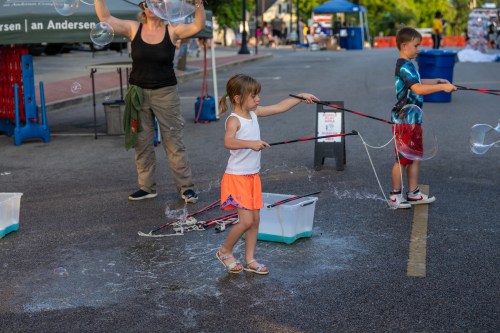 A young girl in a white tank top and orange shorts plays with bubble wands, creating large bubbles on a street.