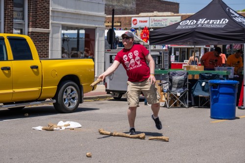 A man in a red shirt throws a stick in a street festival, with a yellow truck and tents in the background.