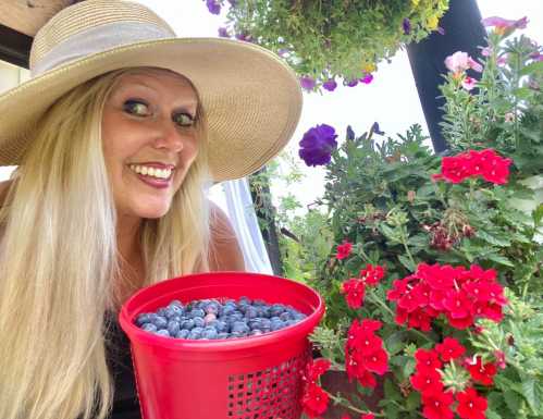 A smiling woman in a wide-brimmed hat holds a basket of blueberries, surrounded by vibrant flowers.