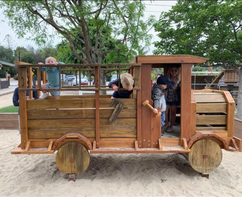 Children playing on a wooden train structure in a playground, surrounded by trees and sand.