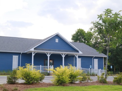A blue building with a porch, surrounded by greenery and shrubs, under a cloudy sky.