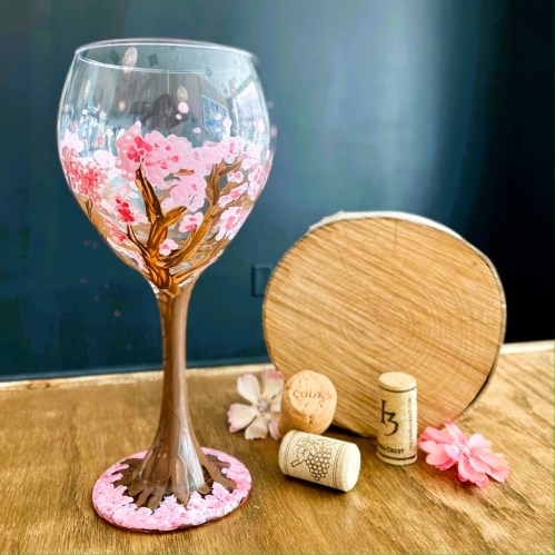 A beautifully painted wine glass featuring pink cherry blossoms, with a wooden coaster and corks in the background.