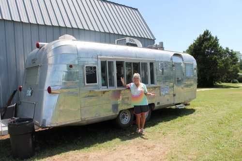 A woman stands in front of a shiny silver food truck parked on grass, with a barn in the background.