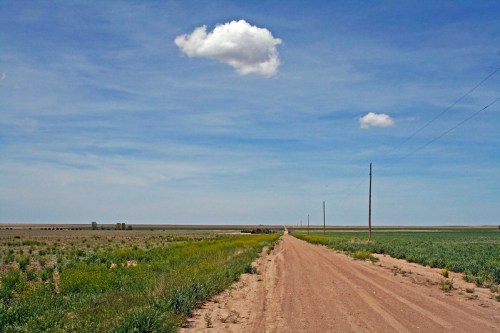 A dirt road stretches through a vast, open landscape with green fields and a blue sky dotted with a single cloud.