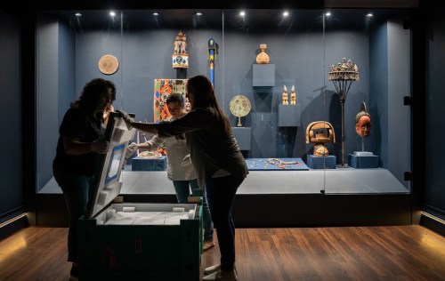 Three people unpacking artifacts in a museum display, surrounded by various cultural items in a well-lit showcase.