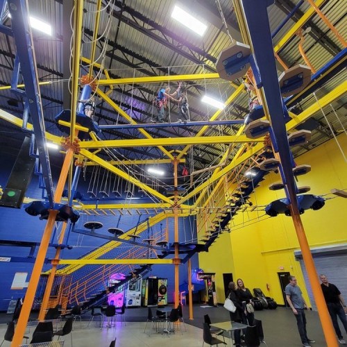 A vibrant indoor adventure park with climbing structures and people navigating ropes high above.
