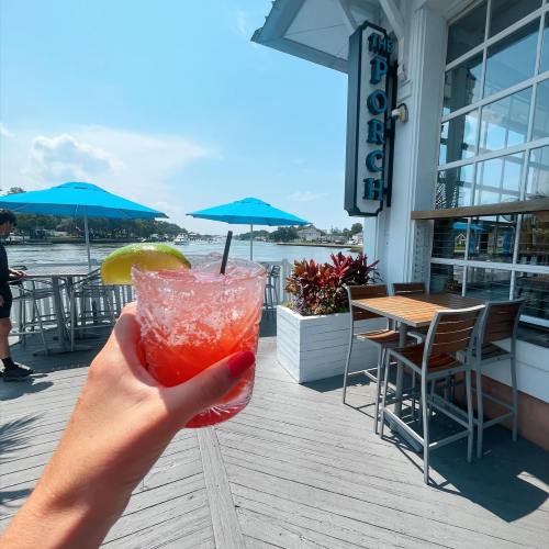 A hand holds a colorful cocktail with a lime slice, with a waterfront restaurant and blue umbrellas in the background.