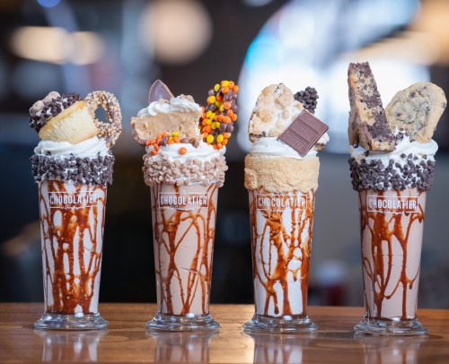 Four decadent milkshakes topped with whipped cream, cookies, and candy, served in tall glasses with chocolate drizzle.