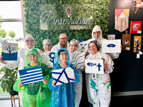 A group of people in protective gear holding painted canvases, smiling in front of a green wall with "Pinspiration" sign.