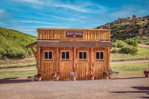 A rustic wooden ticket booth with flower pots, set against a green hillside and blue sky.