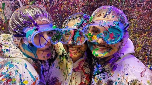 Three people wearing protective gear and goggles, covered in colorful paint splatters, smiling at the camera.