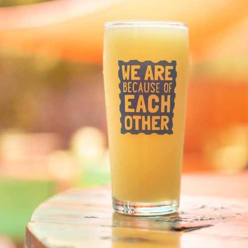 A glass of light-colored beverage with the text "WE ARE BECAUSE OF EACH OTHER" printed on it, set outdoors.