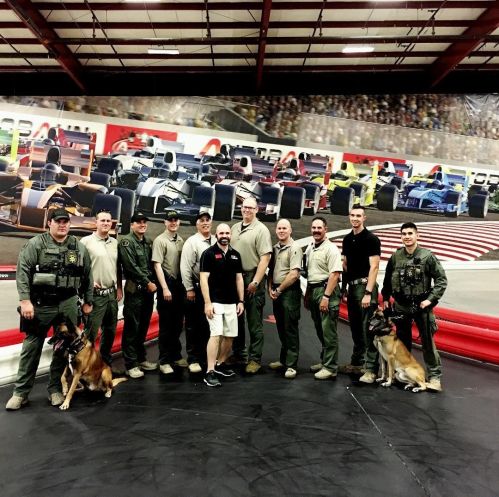 A group of law enforcement officers and their dogs pose together at a racetrack venue.