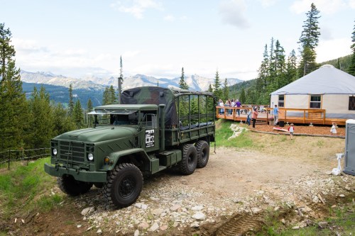 An army green truck parked on a dirt path near a yurt and a group of people enjoying the mountain view.